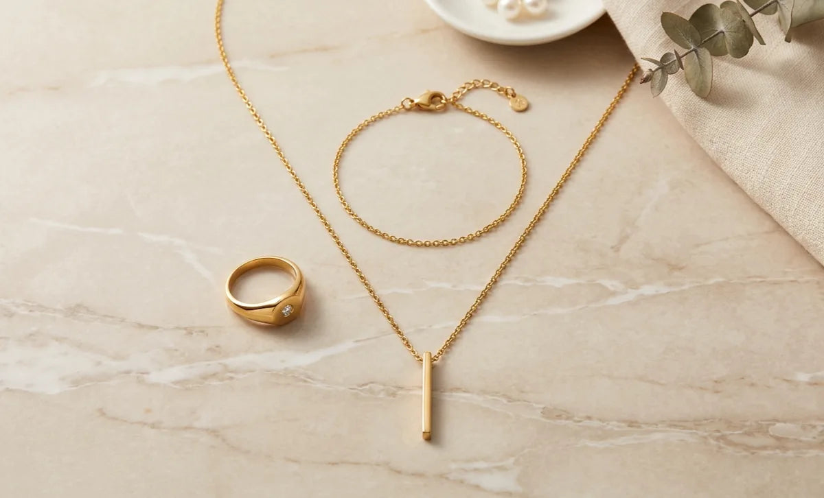 Collier, bracelet et bague dorés en acier inoxydable 316L disposés sur surface beige élégante, collection bijoux ZYORA.