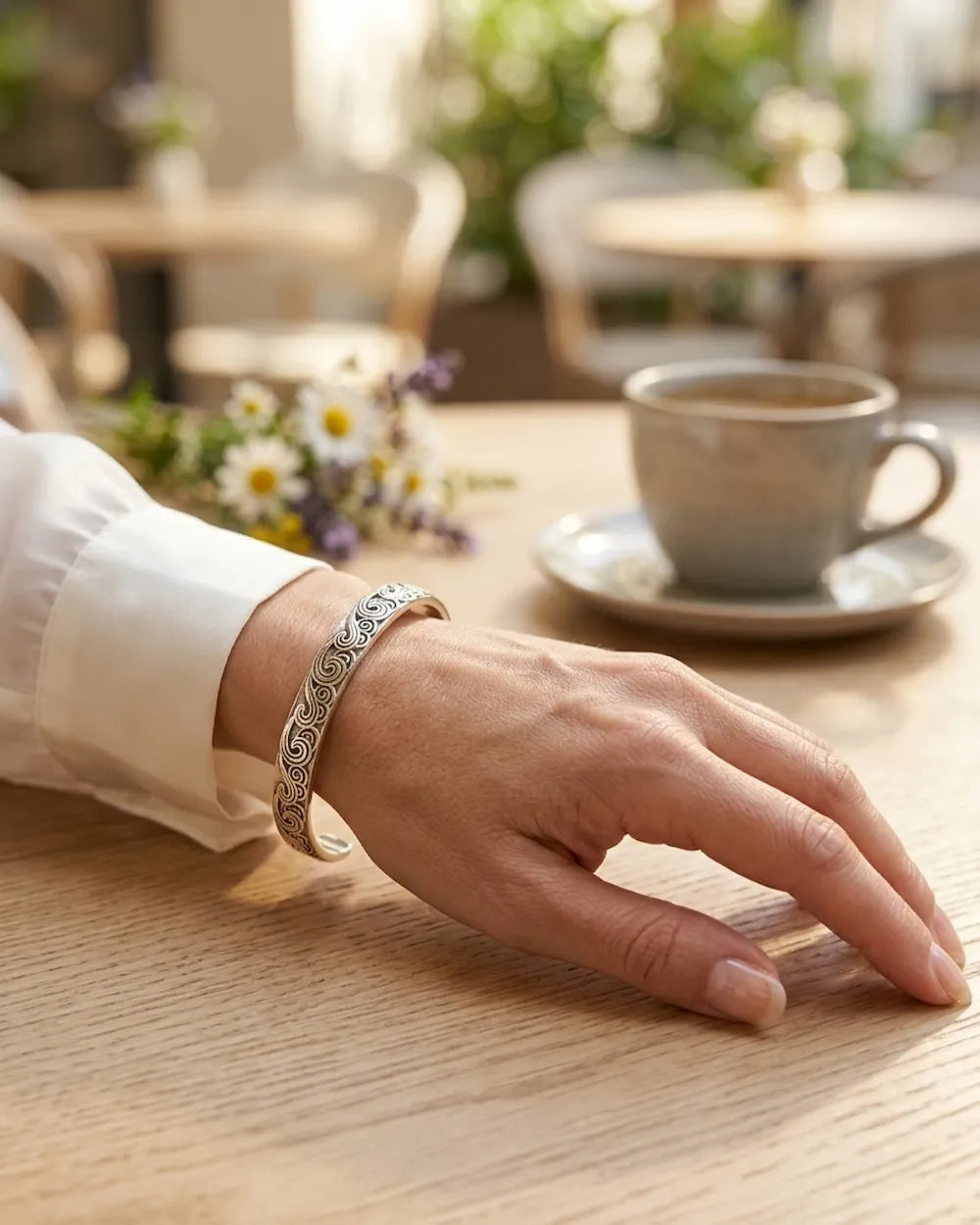 Une photo rapprochée du poignet d'une femme portant un bracelet jonc rigide en argent gravé de motifs complexes de spirales. Sa main reposant sur une table en bois texturée dans un café lumineux. Au premier plan, une tasse à café floue et un petit bouquet de fleurs sauvages. L'ambiance est lumineuse et élégante.